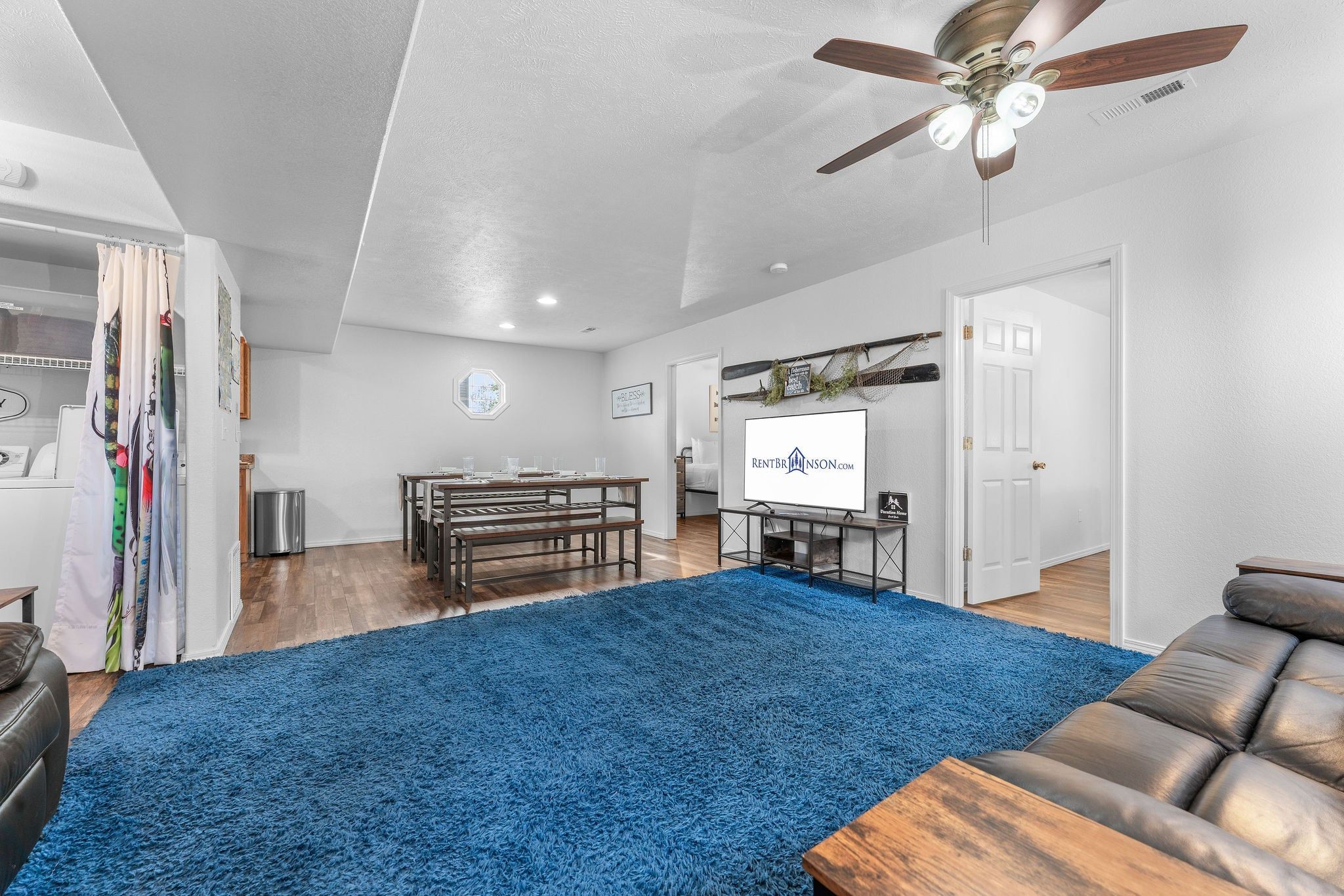 A spacious living room with a blue carpet, a ceiling fan, a wooden dining table, and a sofa.