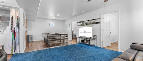 A spacious living room with a blue carpet, a ceiling fan, a wooden dining table, and a sofa.