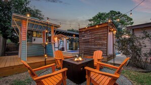 En udendørs terrasse med Adirondack-stole, bålsted og lyskæder.
