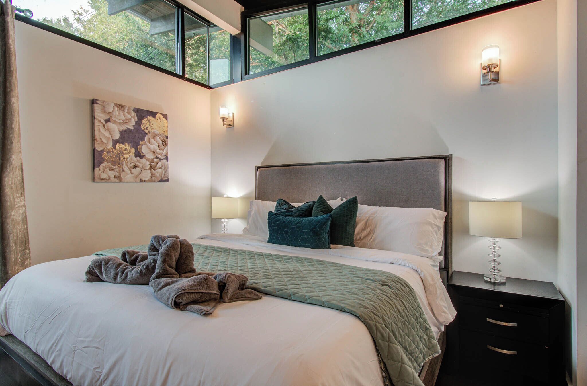 Un dormitorio con una cama grande, dos mesitas de noche con lámparas, un cuadro en la pared y una ventana con vistas a árboles.