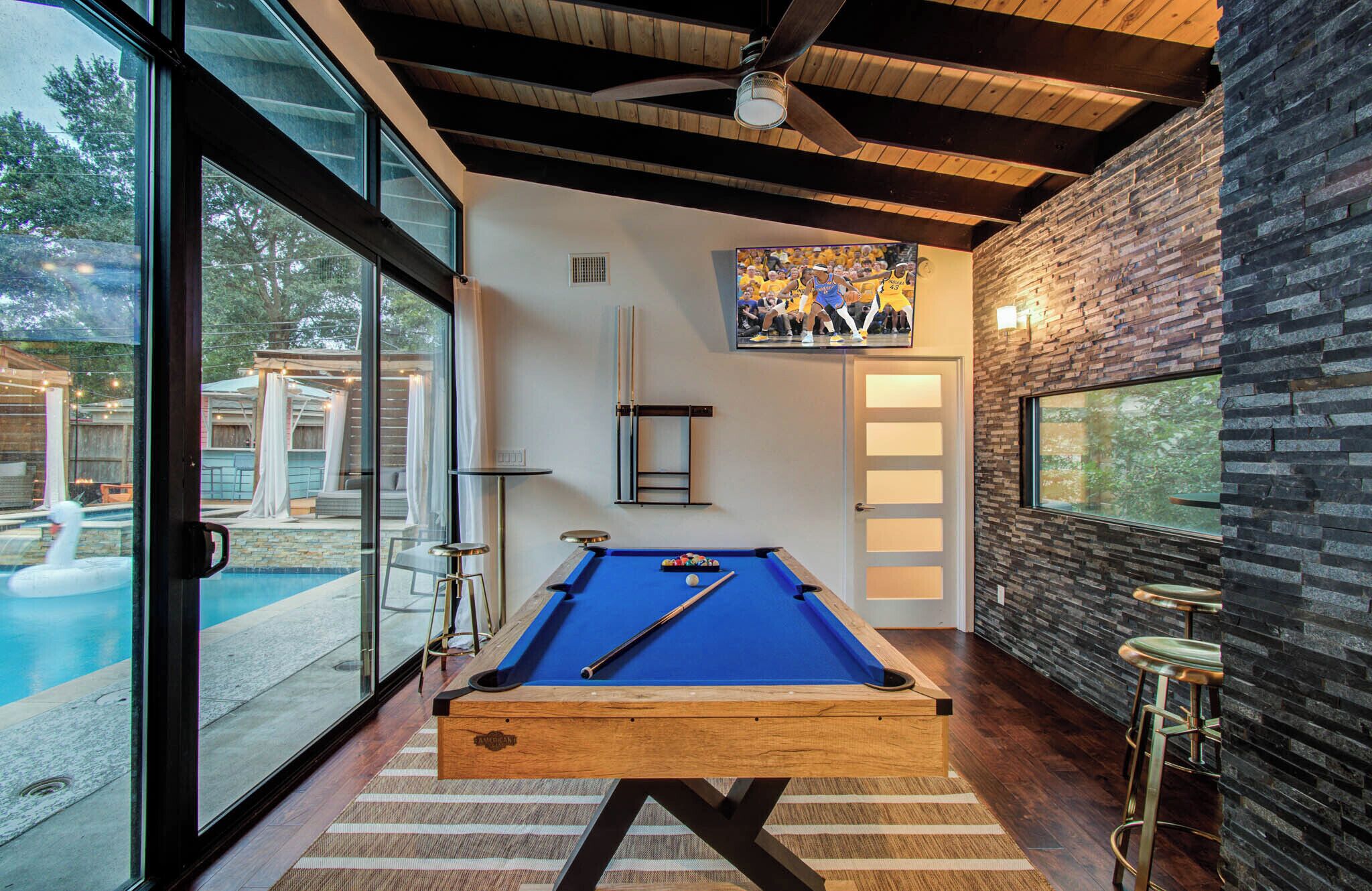 A pool table in a room with a large window, a ceiling fan, and a stone wall.