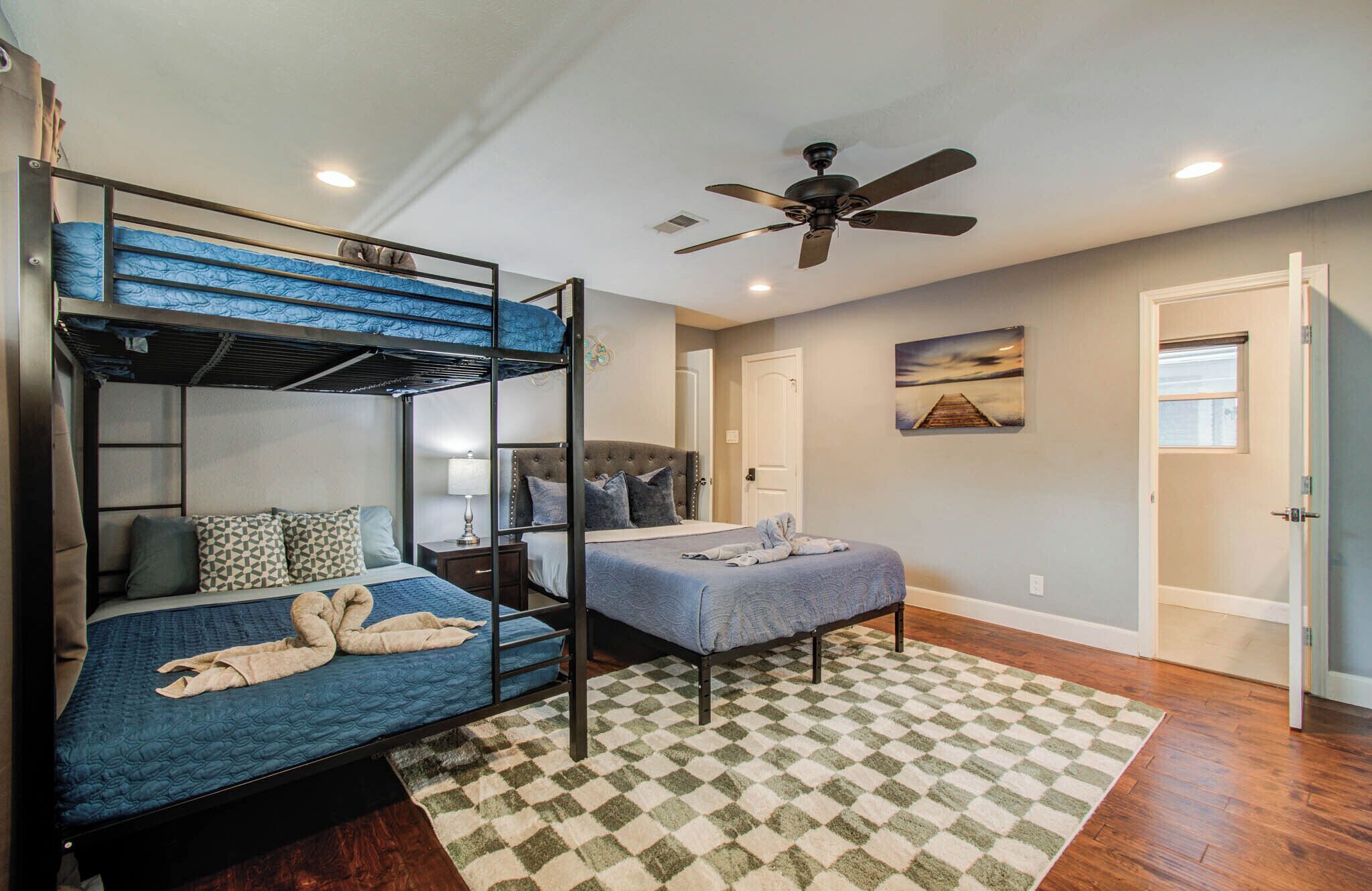 A bunk bed room with a ceiling fan, a bed with a blue quilt, a bedside table with a lamp, and a patterned rug.