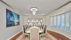 A dining room with a round table, chairs, a chandelier, a large window with blinds, and a framed picture on the wall. - Empress Outdoor Luxury Villa (Houston)