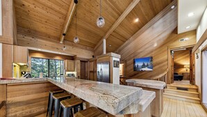 A modern kitchen with wooden accents, a large island with stools, and a view of the outdoors. - Lx16 Luxe Martis Landing Sauna, Gameroom and Hot Tub (Truckee)