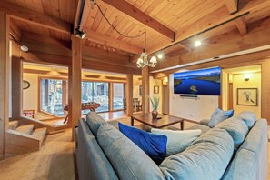 A spacious living area with a large sofa, wooden beams, a chandelier, and a large window offering a view. - Lx16 Luxe Martis Landing Sauna, Gameroom and Hot Tub (Truckee)