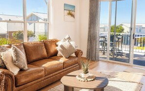 Un sofá de cuero marrón con almohadones decorativos, una mesa redonda de madera con una planta, y vista a un balcón con mobiliario exterior.