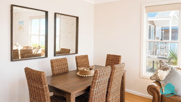 A dining area with a wooden table and wicker chairs, framed artwork on the wall, and a view of a balcony. - Jesbellerai Villa- Caves Beach Resort (Caves Beach)