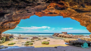 A beach with a natural rock arch, clear blue sky, and calm sea.