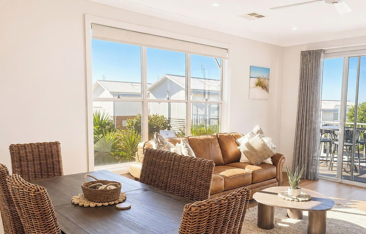 A modern living room with a brown leather sofa, wicker furniture, and a large window offering a view of buildings and greenery.