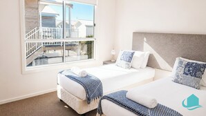 A hotel room with two beds, a large window with a view of buildings, a white headboard, and a small bedside table with a lamp. - Jesbellerai Villa- Caves Beach Resort (Caves Beach)