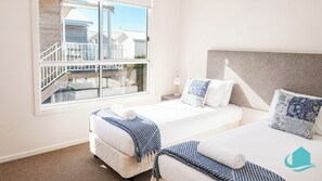 Camera d'albergo con due letti, una grande finestra vista sugli edifici, una testiera bianca e un comodino con una lampada.