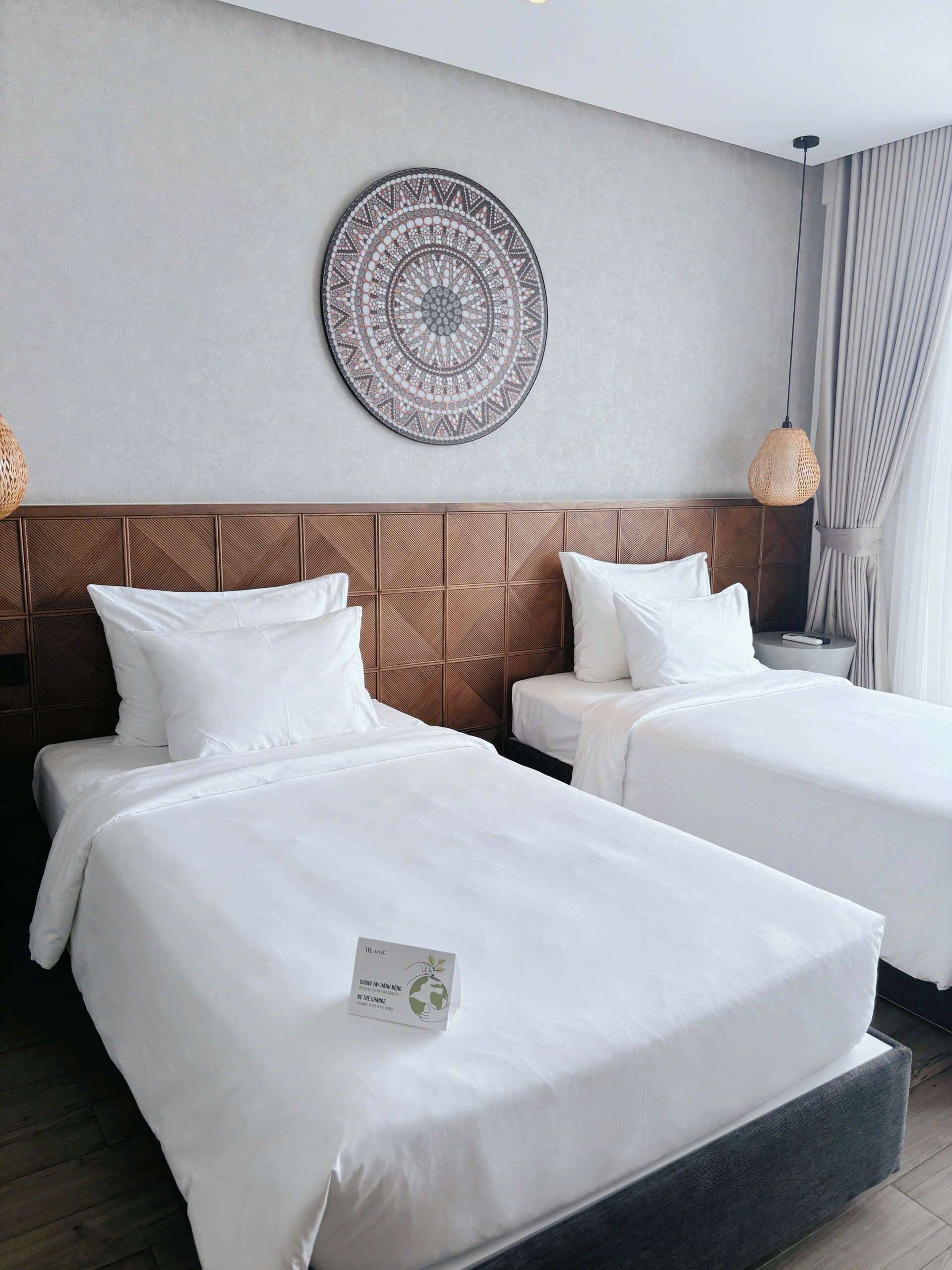 Une chambre d’hôtel avec deux lits, un élément décoratif mural, un luminaire en fibres tressées et une table de chevet avec une télécommande.