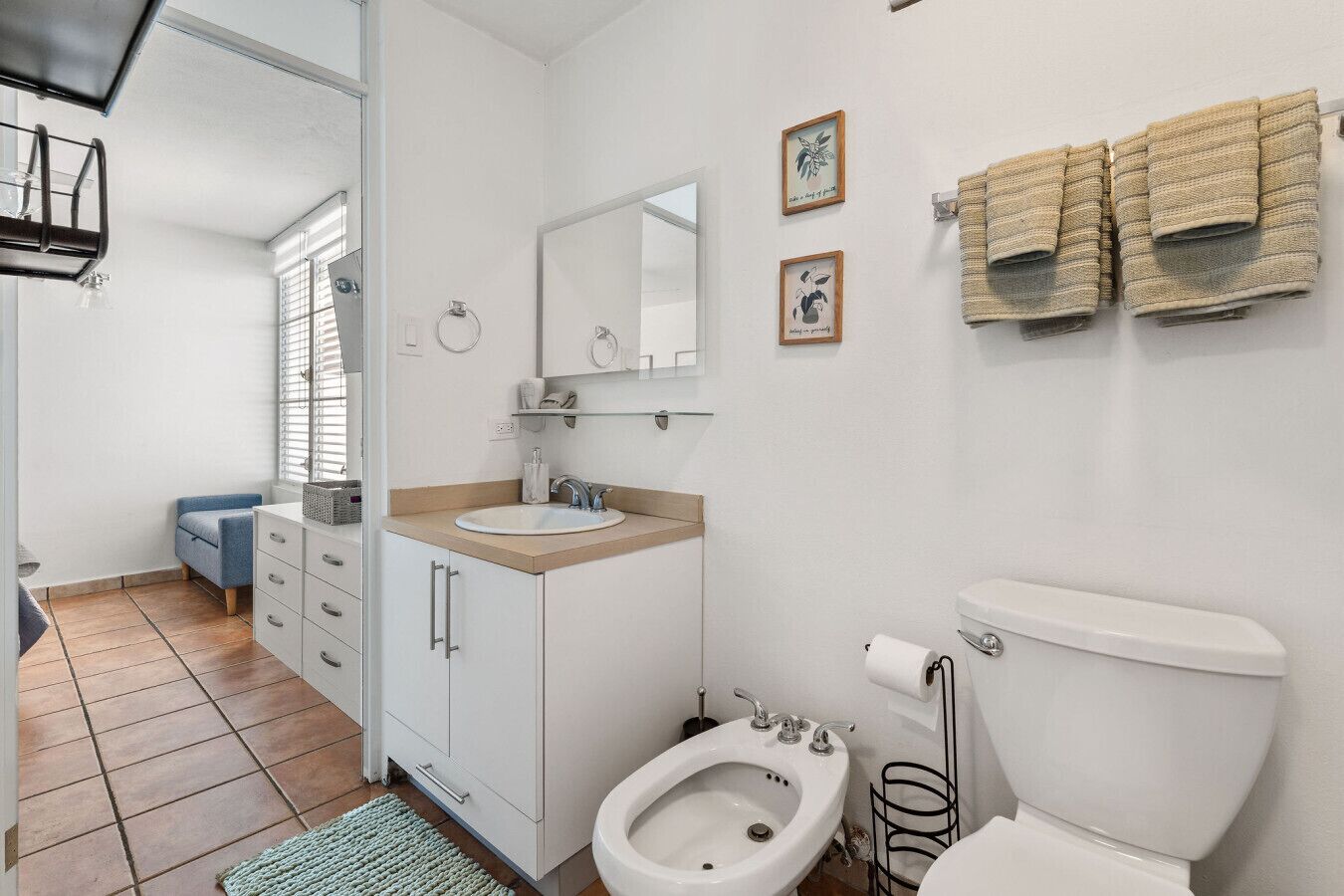 Ein Badezimmer mit Toilette, Bidet, Waschbecken und Spiegel. Handtücher hängen an der Wand und ein kleines Fenster mit Jalousien ist vorhanden.