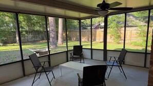 Eine überdachte Terrasse mit vier Stühlen und einem Tisch, großen Fenstern und einem Deckenventilator.