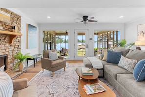 A living room with a stone fireplace, a sofa, a coffee table with books, and a view of a lake through large glass doors. - Best Location On Lake Sinclair (Milledgeville)