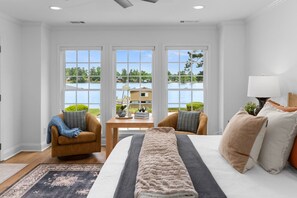 A bedroom with a large bed, two chairs, a table, and a view of a lake and buildings. - Best Location On Lake Sinclair (Milledgeville)