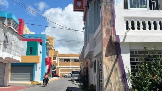 A hotel with a prominent 'HOTEL' sign, a street view with parked cars, and buildings with colorful facades. - Room-hotel Maria Bonita Higüey 2 (Higuey)