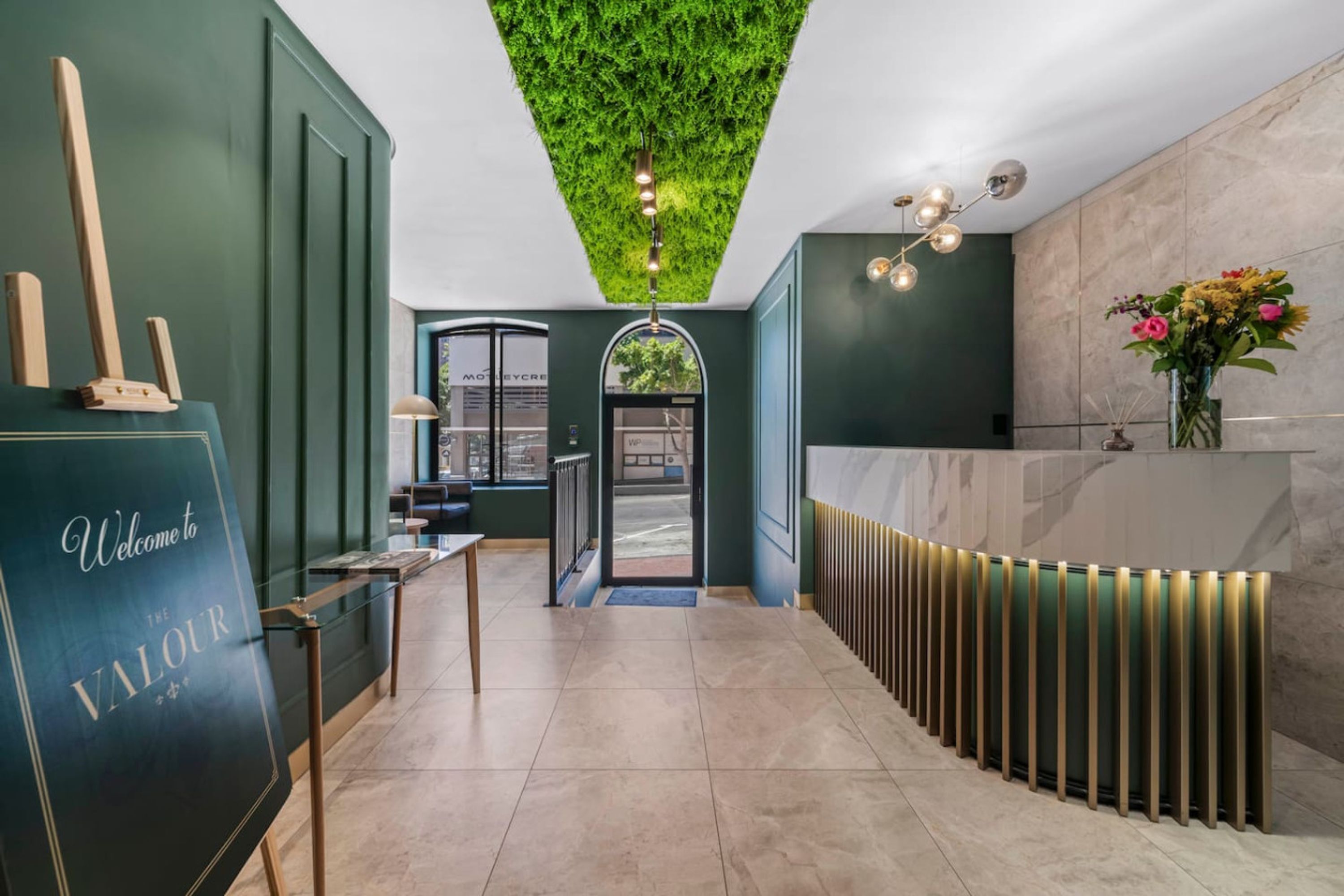 Un espace d’accueil agrémenté d’un mur végétal, d’un comptoir de réception, d’un plafond vert et offrant une vue sur l’extérieur.