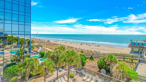A beach view with a boardwalk, palm trees, and a building with a glass facade. - Yachtsman S502 Studio (Myrtle Beach)