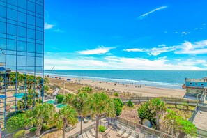 Vista sulla spiaggia con una passerella, palme e un edificio con facciata in vetro.