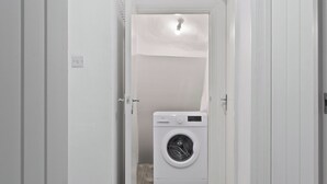 A laundry room with a washing machine, a door leading to another room, and a light fixture on the ceiling. - Cosy 2 Bed-5 Guests-Garden-Parking&WiFi (London)