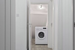 A laundry room with a washing machine, a door leading to another room, and a light fixture on the ceiling. - Cosy 2 Bed-5 Guests-Garden-Parking&WiFi (London)