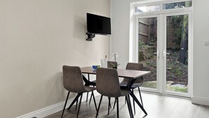 A dining area with a table and chairs, a flat-screen TV mounted on the wall, and a sliding glass door leading to a garden. - Cosy 2 Bed-5 Guests-Garden-Parking&WiFi (London)