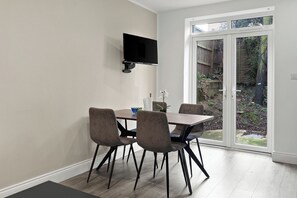A dining area with a table and chairs, a flat-screen TV mounted on the wall, and a sliding glass door leading to a garden. - Cosy 2 Bed-5 Guests-Garden-Parking&WiFi (London)