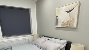 A bedroom with a bed, bedside table, lamp, window, radiator, and artwork on the wall. - Cosy 2 Bed-5 Guests-Garden-Parking&WiFi (London)