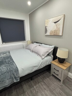 A bedroom with a bed, bedside table, lamp, window, radiator, and artwork on the wall. - Cosy 2 Bed-5 Guests-Garden-Parking&WiFi (London)