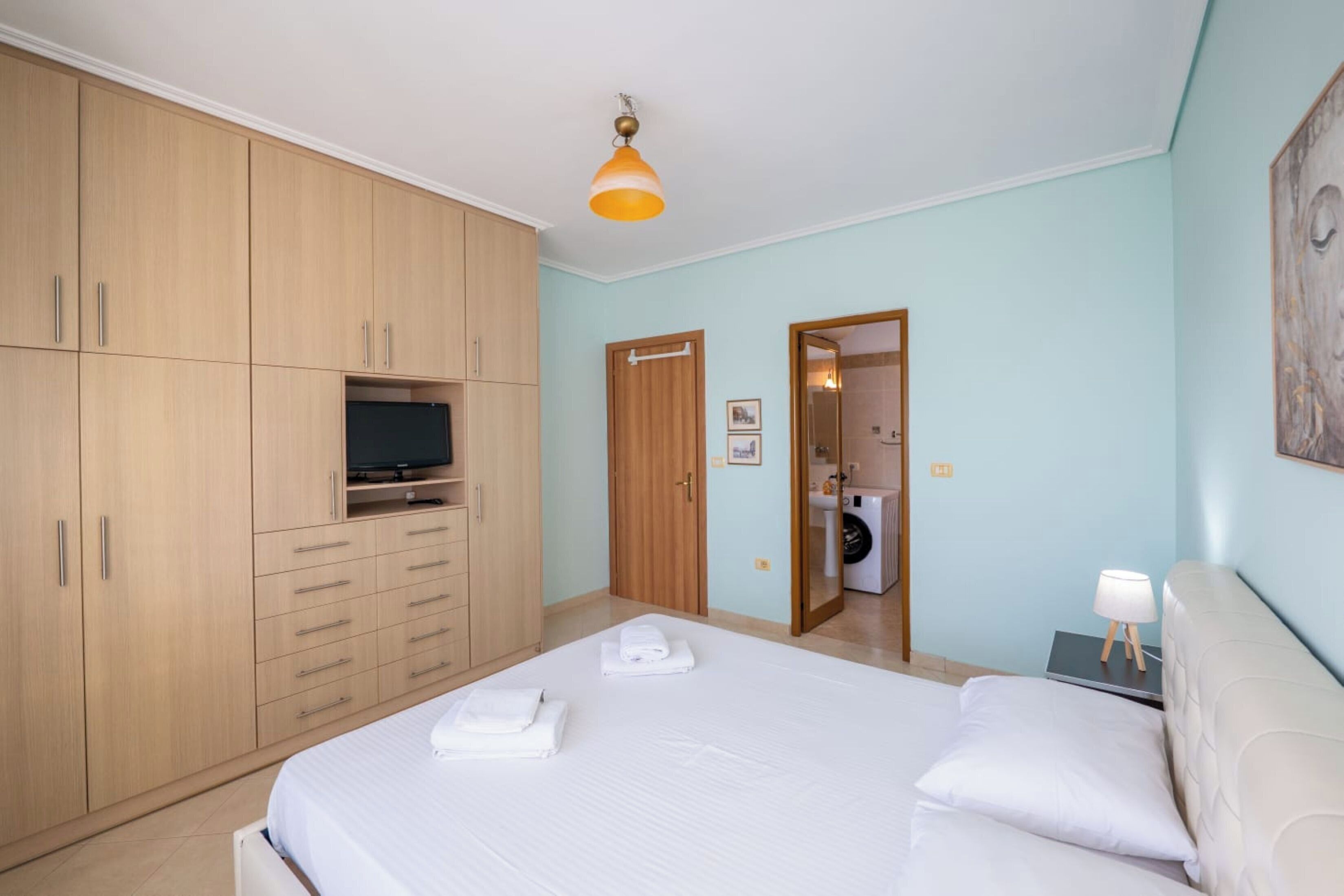 A bedroom with a bed, a TV on a cabinet, a door leading to a bathroom, and a nightstand with a lamp.