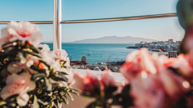Une vue sur la mer, avec une silhouette urbaine en arrière-plan, une balustrade de balcon et quelques fleurs au premier plan.