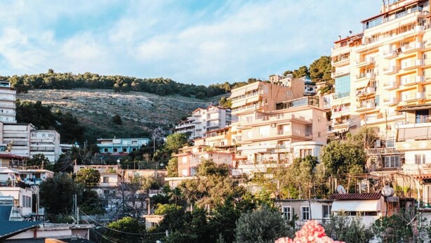 A balcony with a table and chairs, a potted plant, and a view of a hillside with buildings. - Albania Dream Saranda Apartments (Saranda)