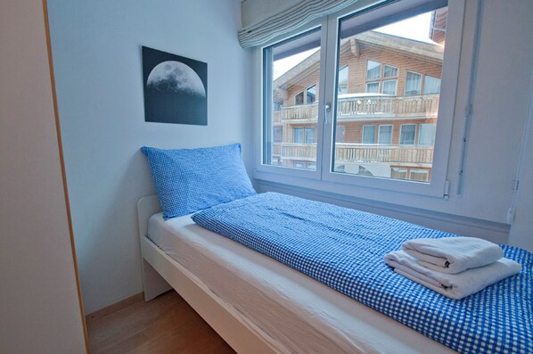 A single bed with a checkered blue and white bedding, a pillow, and a folded towel. A framed picture of a moon is on the wall. - Villa Emeline, Apartment Lora (Zermatt)