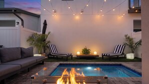 Un jardin avec une piscine, un foyer extérieur, deux chaises longues et des guirlandes lumineuses.
