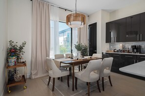 Una cocina moderna con armarios oscuros, un comedor con mesa de madera y sillas blancas, y un amplio ventanal con cortinas translúcidas.
