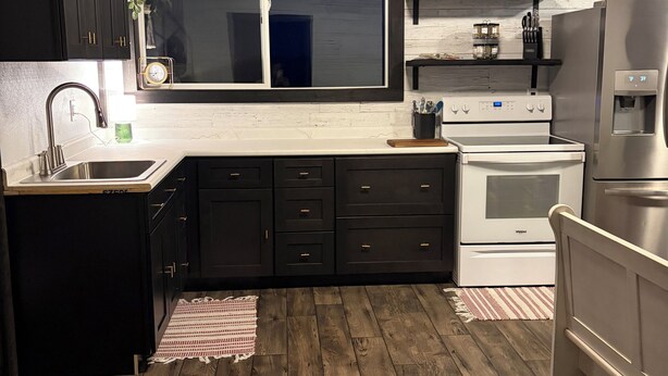 A modern kitchen with dark cabinets, a stainless steel refrigerator, a built-in oven, and a window above the sink.