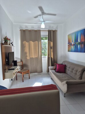 A living room with a ceiling fan, a sofa, a chair, a TV, and a painting on the wall. - Apartment in the best location in Praia do Forte. Beachfront property with garage. (Cabo Frio)