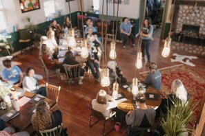 Una habitación amplia con suelo de madera, lámparas colgantes y un grupo de personas sentadas en mesas.