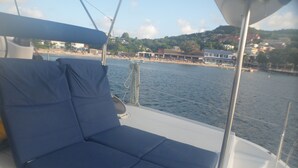 A boat deck with a blue cushioned seating area, metal railings, and a view of a coastal town with buildings and a beach. - Shared catamaran anchored in anse mitan bay at trois ilets (Les Trois-Îlets)