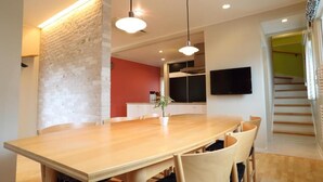 A modern dining area with a wooden table, chairs, pendant lighting, a brick accent wall, a kitchen area with cabinets, a flat-screen TV, and a staircase. - Tabinoteitaku Kamakura Yuigahama (Kamakura)
