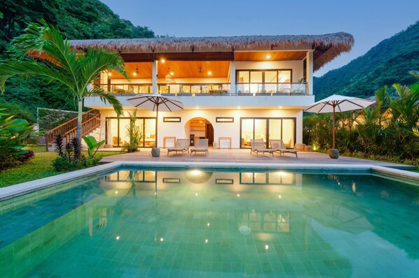 A two-story house with a pool, surrounded by lush greenery and hills.