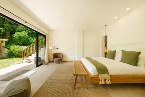 A modern bedroom with a large bed, a chair, a small table, and a view of a garden through a sliding door.