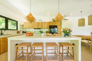 A modern kitchen with wooden cabinets, a central island with stools, hanging wicker lights, and a view of greenery outside.