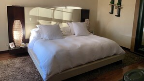 Un lit bien fait, avec du linge de lit blanc, une tête de lit moderne, une lampe de sol décorative et une photo encadrée posée sur une table de chevet.