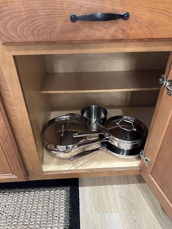 Un mueble de madera con un tirador metálico, que contiene utensilios de cocina de acero inoxidable, incluyendo una olla y sartenes.