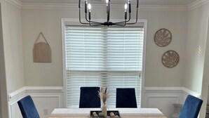 Un coin repas agrémenté d’un lustre, d’une table recouverte d’une nappe blanche, de chaises à l’assise bleue et d’une fenêtre avec des stores.