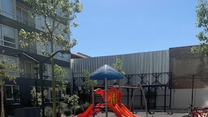 Ein Spielplatz mit Rutsche und Klettergerüsten, umgeben von einem befestigten Bereich mit taktilen Bodenindikatoren für Barrierefreiheit.