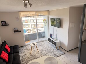 A living room with a black sofa, a small round table, a TV mounted on the wall, and a balcony with a view of buildings.
