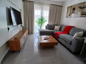 Ein modernes Wohnzimmer mit grauem Sofa, einem Holztisch, einem flachbildfernseher an der Wand und einem großen Fenster mit Vorhängen.